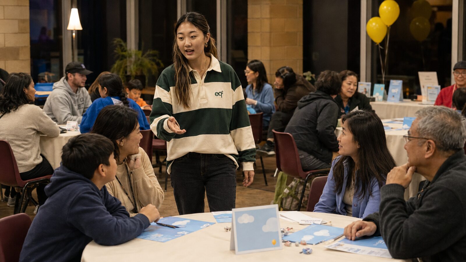 Emma Byun at an evening community event with round tables and balloons, talking with a multi-generational family group